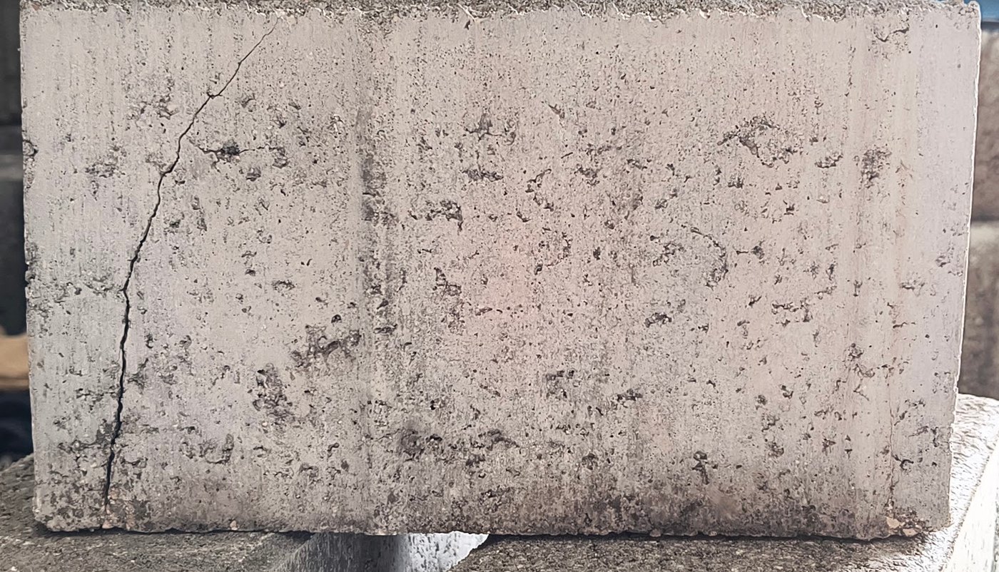 Adoquín de concreto de 120 mm roto en el ensayo de flexotracción, visto de lado: la grieta nace en la cara inferior junto al extremo izquierdo, sube casi vertical y se inclina hacia el centro al llegar arriba