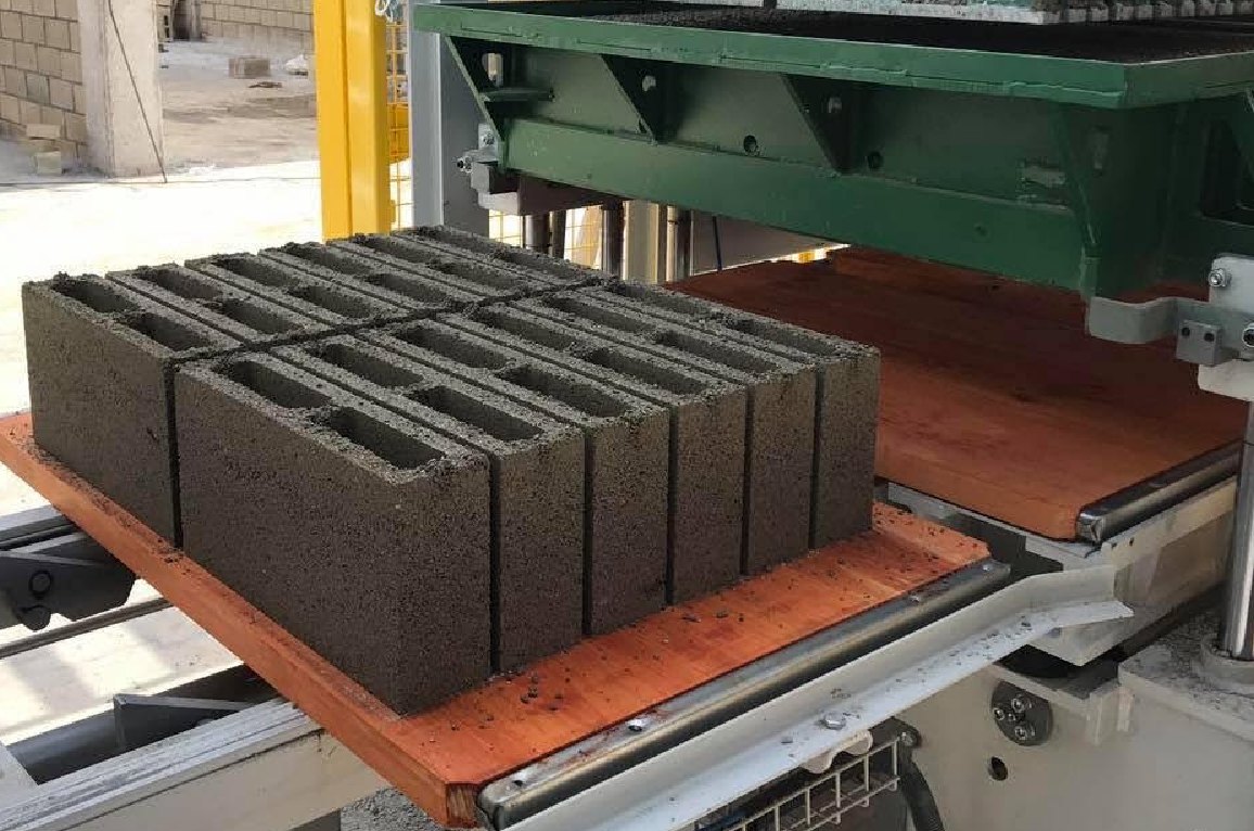 Bloques de concreto recién prensados sobre una bandeja de madera, saliendo de la vibroprensa hacia el transportador