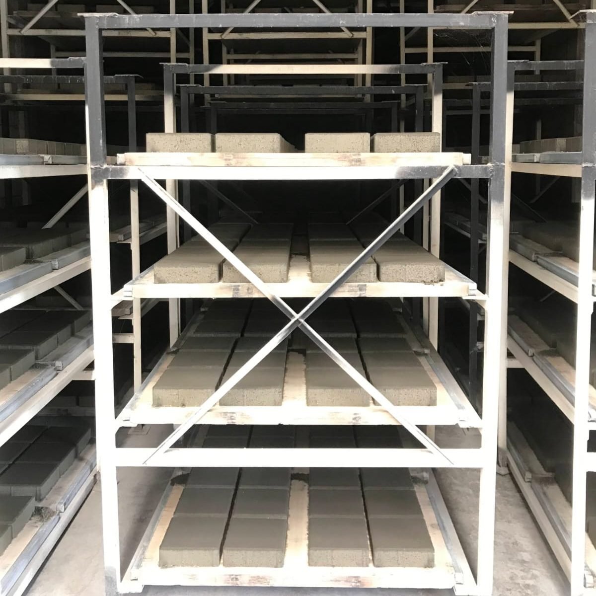 Estantes metálicos de curado cargados de bandejas de madera con adoquines recién moldeados, cuatro niveles en cada estante, y detrás y a los lados más estantes llenos de bandejas: cada bandeja de un día de producción necesita su lugar bajo techo