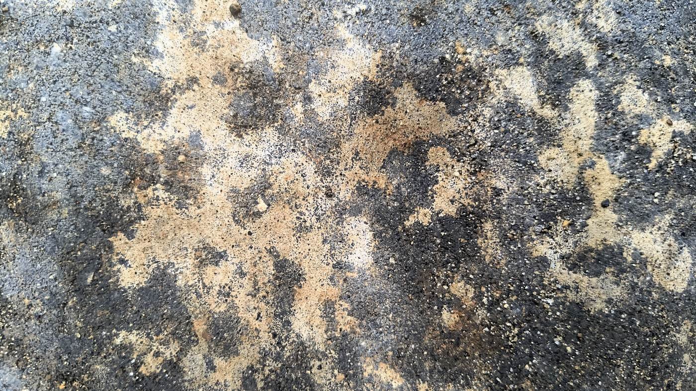 Detalle de una pieza de concreto oscura con un depósito beige granular y moteado sobre la cara vista, del mismo color que el material fino del entorno