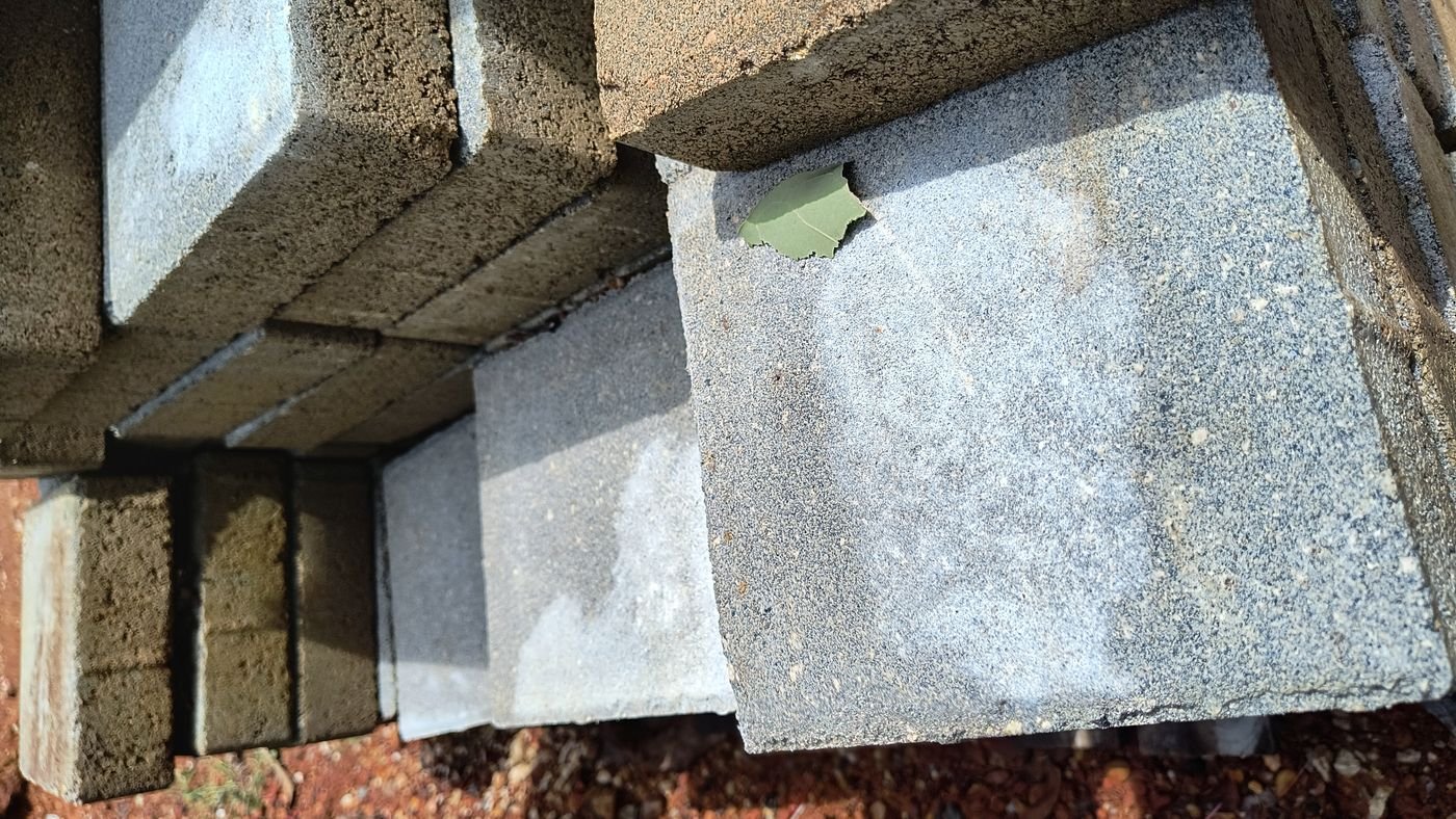 Piezas de concreto apiladas a la intemperie en el patio de una obra, sobre el terreno y sin cubrir, con un velo blanquecino uniforme sobre la cara vista de las de arriba