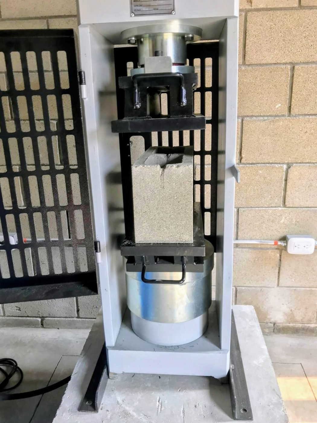 Máquina de ensayo de compresión con platos para mampostería: un bloque de concreto entre dos platos de acero gruesos y rectangulares que lo cubren por completo, arriba y abajo, con la rótula sobre el plato superior